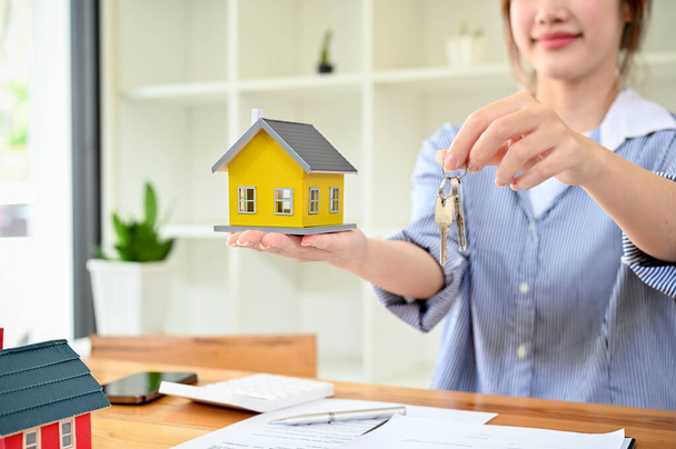 Charming asian female real estate agent holding a beautiful house model and house keys over the office desk. Property Real estate purchasing concept. - Foto, Bild