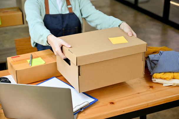 Cropped image, Female seller holding a big cardboard box or brown parcel box over her desk, packing the customer's order. Fashion shop owner. - Photo, Image