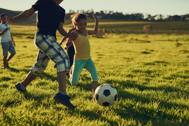 Fun with friends and a football. Shot of a group of children playing soccer together in a field outside. - 写真・画像