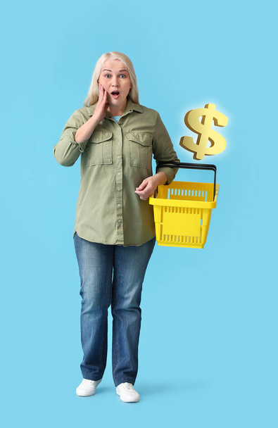 Shocked mature woman with empty shopping basket on blue background. Price increase - Photo, Image