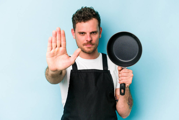 Молода кавказька плита тримає сковороду ізольовано на синьому фоні, що стоїть з розтягнутою рукою, що показує знак стоп, запобігаючи вам
. - Фото, зображення