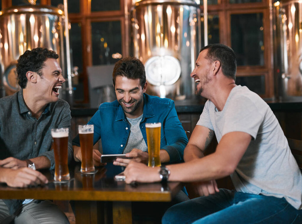 Time to talk business over a beer. Shot of three cheerful young men drinking beer together at a table inside of a beer brewery during the day. - 写真・画像