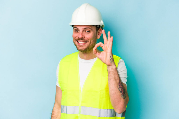 Young laborer caucasian man isolated on blue background cheerful and confident showing ok gesture. - Photo, image