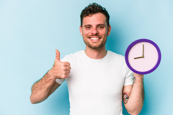 Young caucasian man holding a clock isolated on blue background smiling and raising thumb up - Foto, immagini