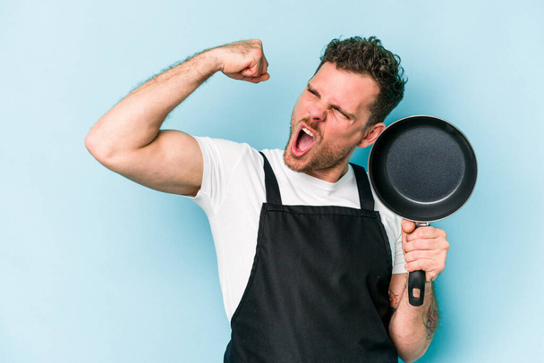 Молода кавказька плита тримає сковороду ізольовано на синьому фоні, піднімаючи кулак після перемоги, концепція переможця
. - Фото, зображення