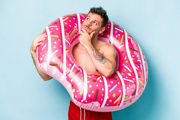 Young caucasian man holding an inflatable donut isolated on blue background looking sideways with doubtful and skeptical expression. - Photo, image