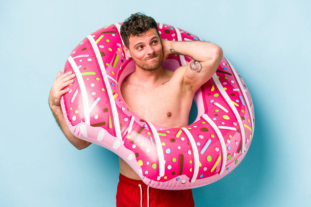 Young caucasian man holding an inflatable donut isolated on blue background touching back of head, thinking and making a choice. - Valokuva, kuva