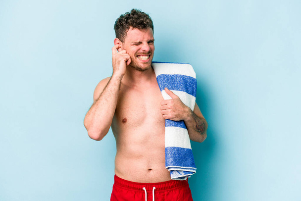 Jovem caucasiano segurando toalha de praia isolada no fundo azul cobrindo orelhas com as mãos. - Foto, Imagem