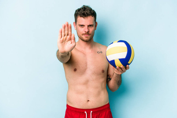 Молодий білий чоловік грає волейбол ізольовано на синьому фоні, стоїть з простягнутою рукою, показуючи знак зупинки, запобігаючи вам
. - Фото, зображення