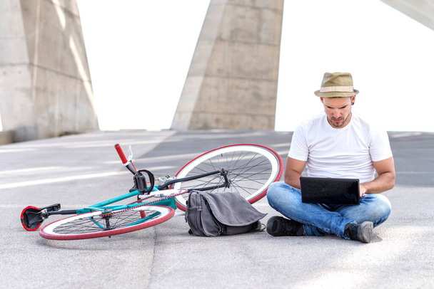 Portret młodego mężczyzny, siedzącego na ziemi z skrzyżowanymi nogami i pracującego na laptopie podczas odpoczynku - Zdjęcie, obraz
