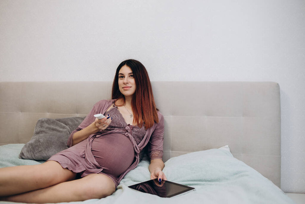 side view on relaxed pregnant lady watching movies on tv, brunette woman in domestic wear sits on sofa with plate with popcorn - Foto, imagen