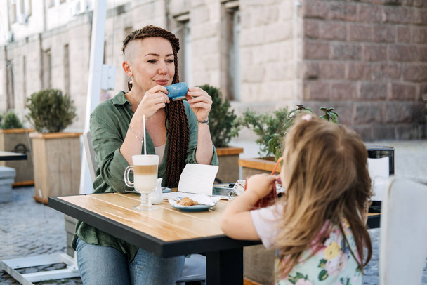 Mother and little daughter eating in the street cafe. Trendy hipster Mother with dreadlocks and toddler daughter having lunch together at cafe summer day - Foto, Imagem