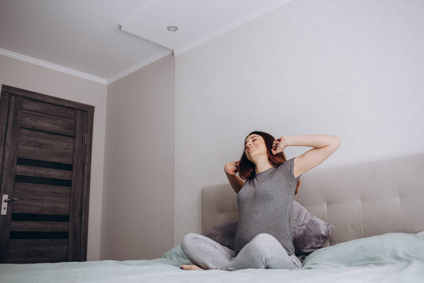 беременная женщина растягивается утром, сидя на кровати, молодая женщина в пижаме просыпается - Фото, изображение