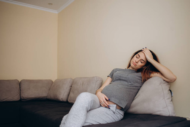 Jovem bela mulher grávida sentada em casa no sofá doente, tem dor de cabeça e náusea - Foto, Imagem