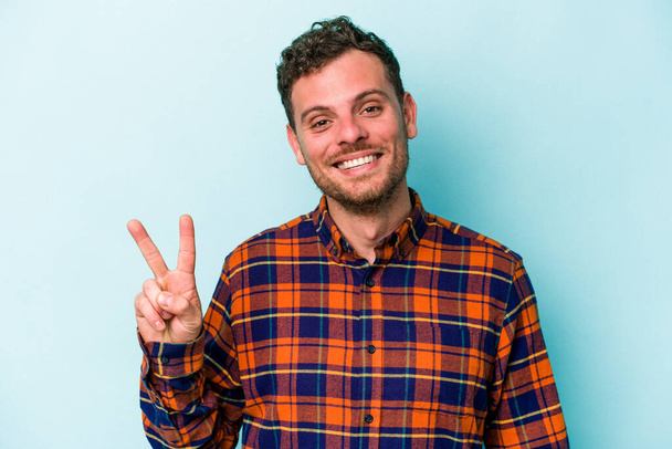 Young caucasian man isolated on blue background showing victory sign and smiling broadly. - Photo, Image