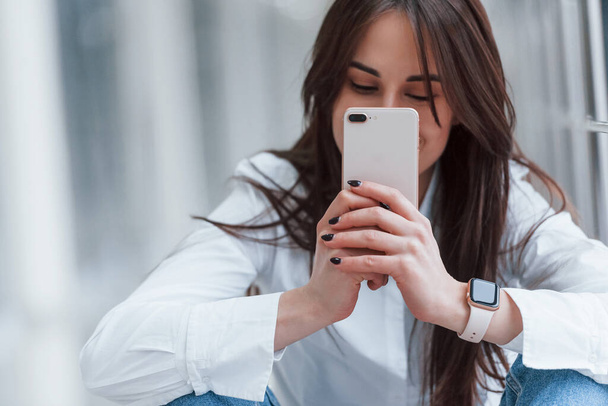 Brunette in white shirt with phone in hands sits indoors in modern airport or hallway at daytime. - Foto, imagen