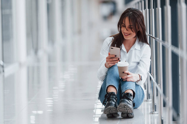 Brunette in white shirt sits indoors in modern airport or hallway at daytime with phone and cup of drink. - Fotografie, Obrázek