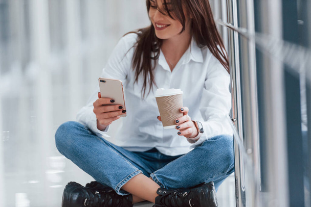 Brunette in white shirt sits indoors in modern airport or hallway at daytime with phone and cup of drink. - Foto, Imagen