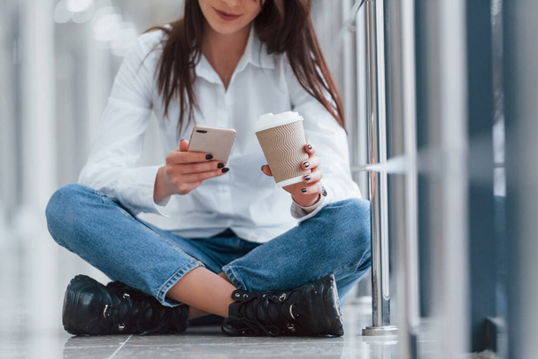 Brunette in white shirt sits indoors in modern airport or hallway at daytime with phone and cup of drink. - Foto, immagini