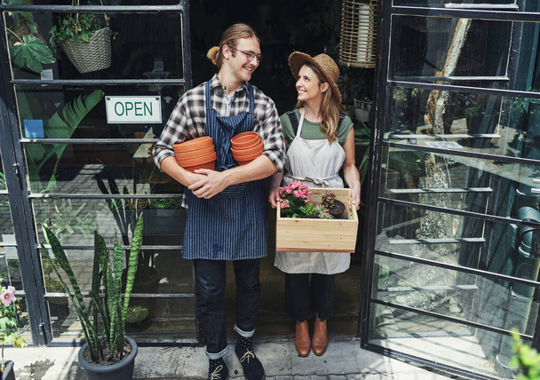 Tiimityöllä pääsee pitkälle. Täyspitkä laukaus kahdesta nuoresta yrittäjästä, jotka seisovat kukkakaupan sisäänkäynnin vieressä yhdessä ja pitävät ruukkutarvikkeita. - Valokuva, kuva