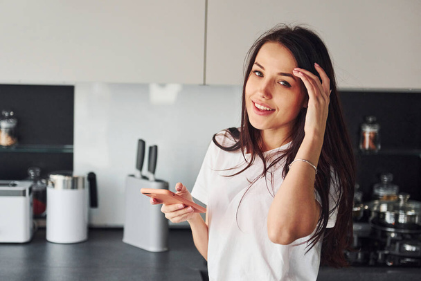 Young beautiful brunette in casual clothes indoors in kitchen at daytime using smartphone. - Foto, Imagem