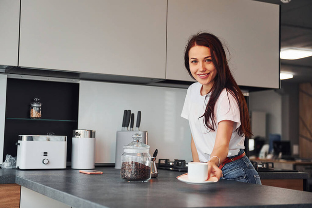 Gives fresh coffee. Young beautiful brunette in casual clothes indoors in kitchen at daytime. - Fotoğraf, Görsel