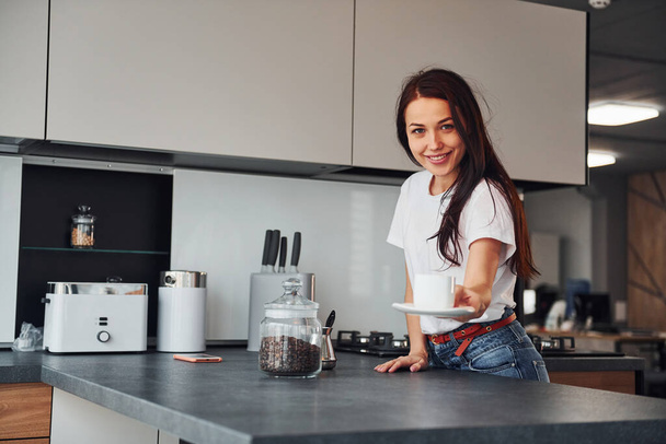 Gives fresh coffee. Young beautiful brunette in casual clothes indoors in kitchen at daytime. - Фото, зображення