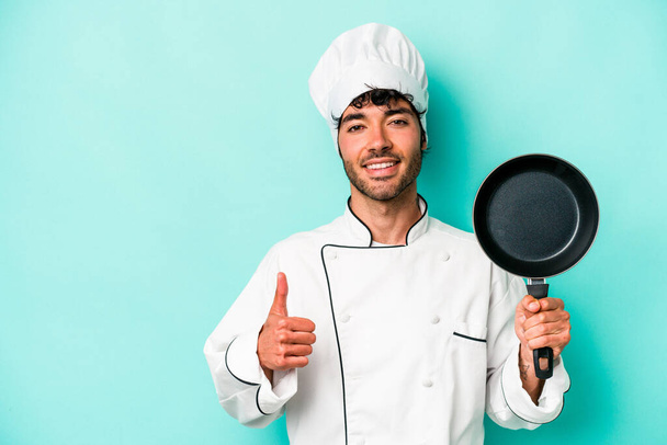 Young caucasian chef man holding flying pan isolated on blue background smiling and raising thumb up - Foto, immagini