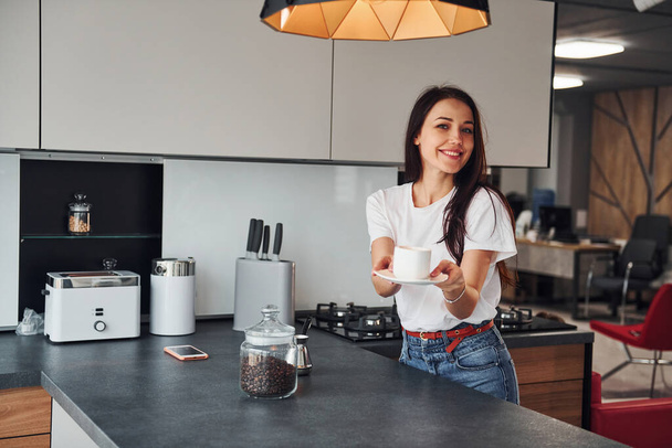 Gives fresh coffee. Young beautiful brunette in casual clothes indoors in kitchen at daytime. - Foto, afbeelding