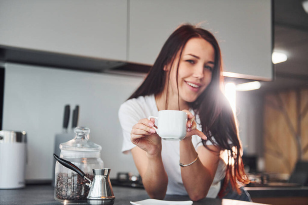With cup of fresh coffee. Young beautiful brunette in casual clothes indoors in kitchen at daytime. - Фото, изображение