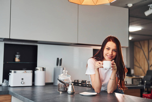With cup of fresh coffee. Young beautiful brunette in casual clothes indoors in kitchen at daytime. - 写真・画像