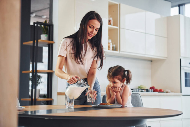 Mother pouring fresh milk into the glass for her little daughter in kitchen. - Fotoğraf, Görsel