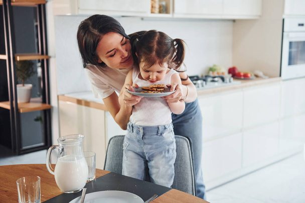 Little girl eats cookies. Mother with her little daughter indoors in kitchen. - Zdjęcie, obraz