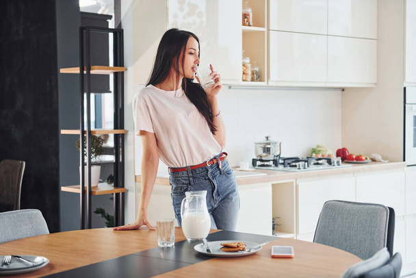 Young beautiful brunette in casual clothes indoors in kitchen at daytime near table with milk and cookies. - Φωτογραφία, εικόνα