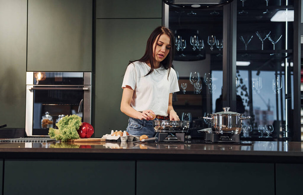 Preparing food by using vegetables and healthy products. Young beautiful brunette in casual clothes indoors in kitchen at daytime. - Фото, зображення
