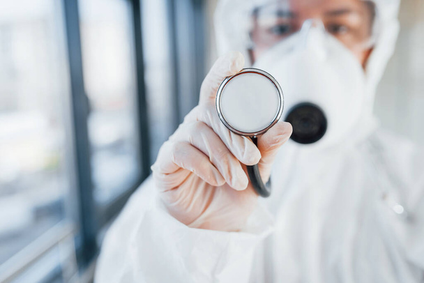 Female doctor scientist in lab coat, defensive eyewear and mask standing indoors and holds stethoscope. - Foto, Imagem