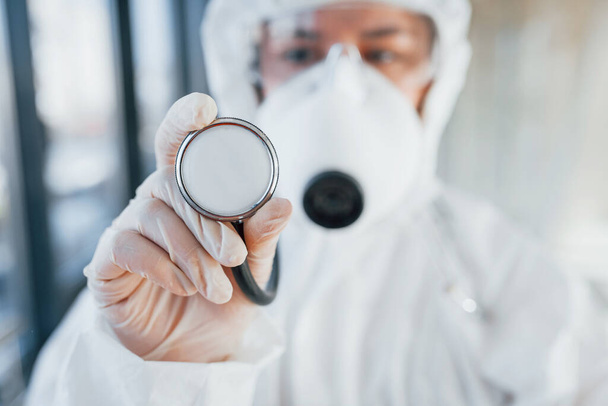 Female doctor scientist in lab coat, defensive eyewear and mask standing indoors and holds stethoscope. - Foto, Bild