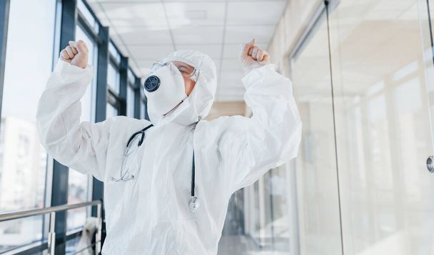 Celebrating victory. Female doctor scientist in lab coat, defensive eyewear and mask standing indoors. - Foto, Imagem