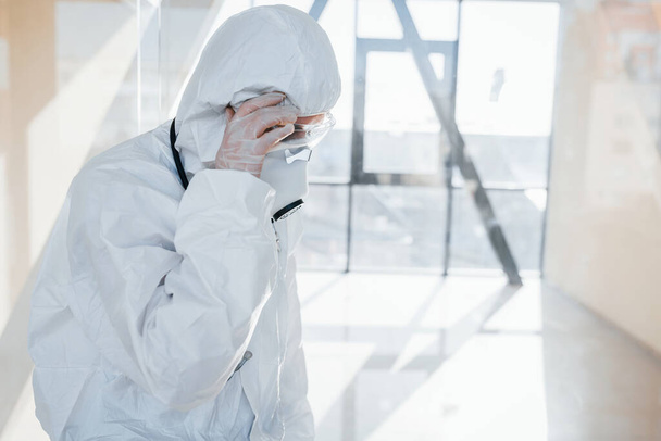 Feels bad and sick. Female doctor scientist in lab coat, defensive eyewear and mask standing indoors. - Фото, зображення