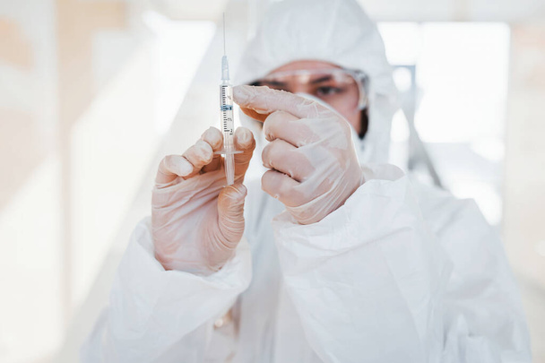 Holds syringe with medicine. Female doctor scientist in lab coat, defensive eyewear and mask standing indoors. - Foto, Imagen