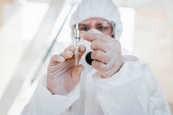 Holds syringe with medicine. Female doctor scientist in lab coat, defensive eyewear and mask standing indoors. - Foto, Imagem