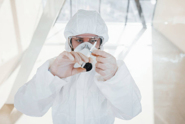 Holds syringe with medicine. Female doctor scientist in lab coat, defensive eyewear and mask standing indoors. - Photo, Image