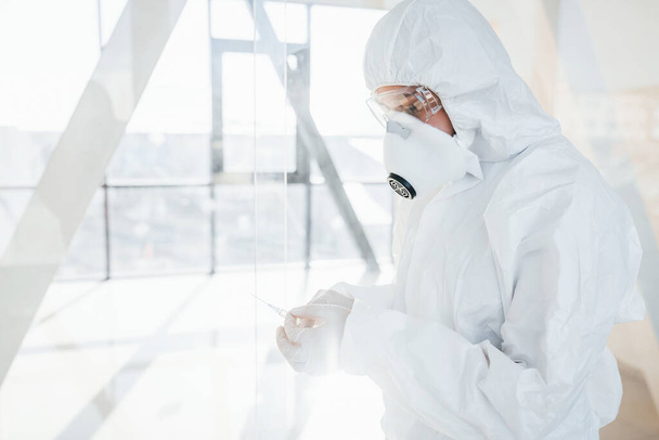 Holds syringe with medicine. Female doctor scientist in lab coat, defensive eyewear and mask standing indoors. - Photo, Image