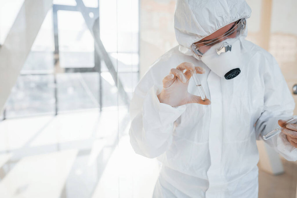 With test tube in hand. Female doctor scientist in lab coat, defensive eyewear and mask standing indoors. - Photo, Image