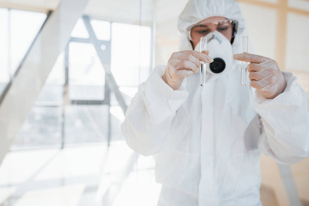 With test tube in hand. Female doctor scientist in lab coat, defensive eyewear and mask standing indoors. - Photo, Image