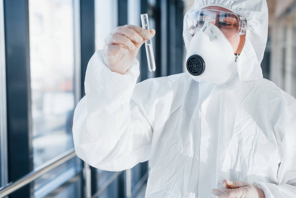 With test tube in hand. Female doctor scientist in lab coat, defensive eyewear and mask standing indoors. - Fotó, kép