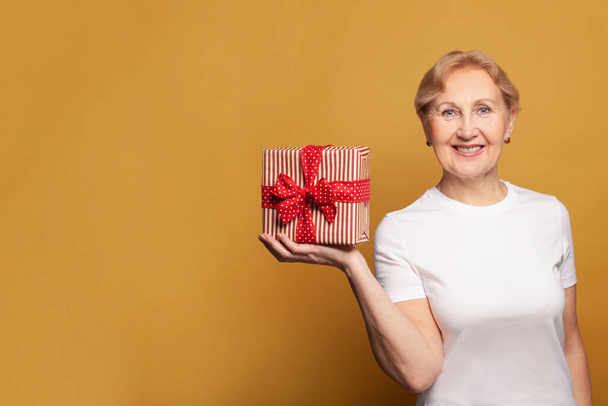 Ελκυστική χαμογελαστή ηλικιωμένη γυναίκα σε λευκό t-shirt κρατώντας κόκκινο κουτί δώρου σε φωτεινό κίτρινο φόντο banner τοίχο στούντιο - Φωτογραφία, εικόνα