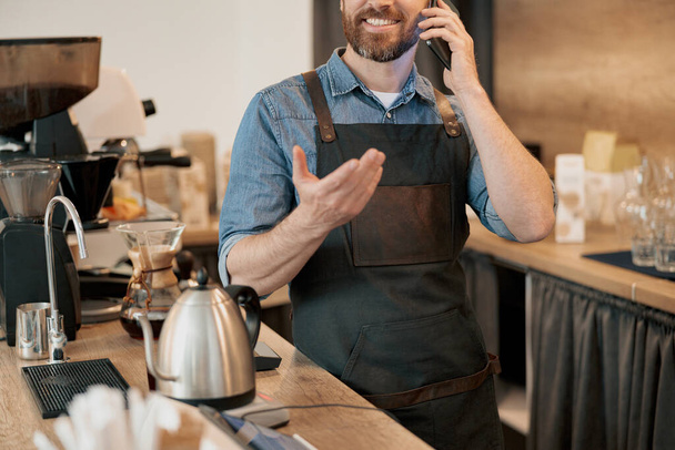 Sonriente barista habla de teléfono de pie detrás del mostrador en la cafetería - Foto, imagen