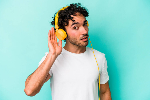 Giovane uomo caucasico ascoltare musica isolata su sfondo blu cercando di ascoltare un pettegolezzo. - Foto, immagini