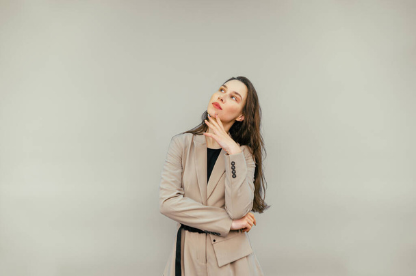 Pensive business lady in a jacket stands on a beige background and looks away and thinks. - Foto, Bild